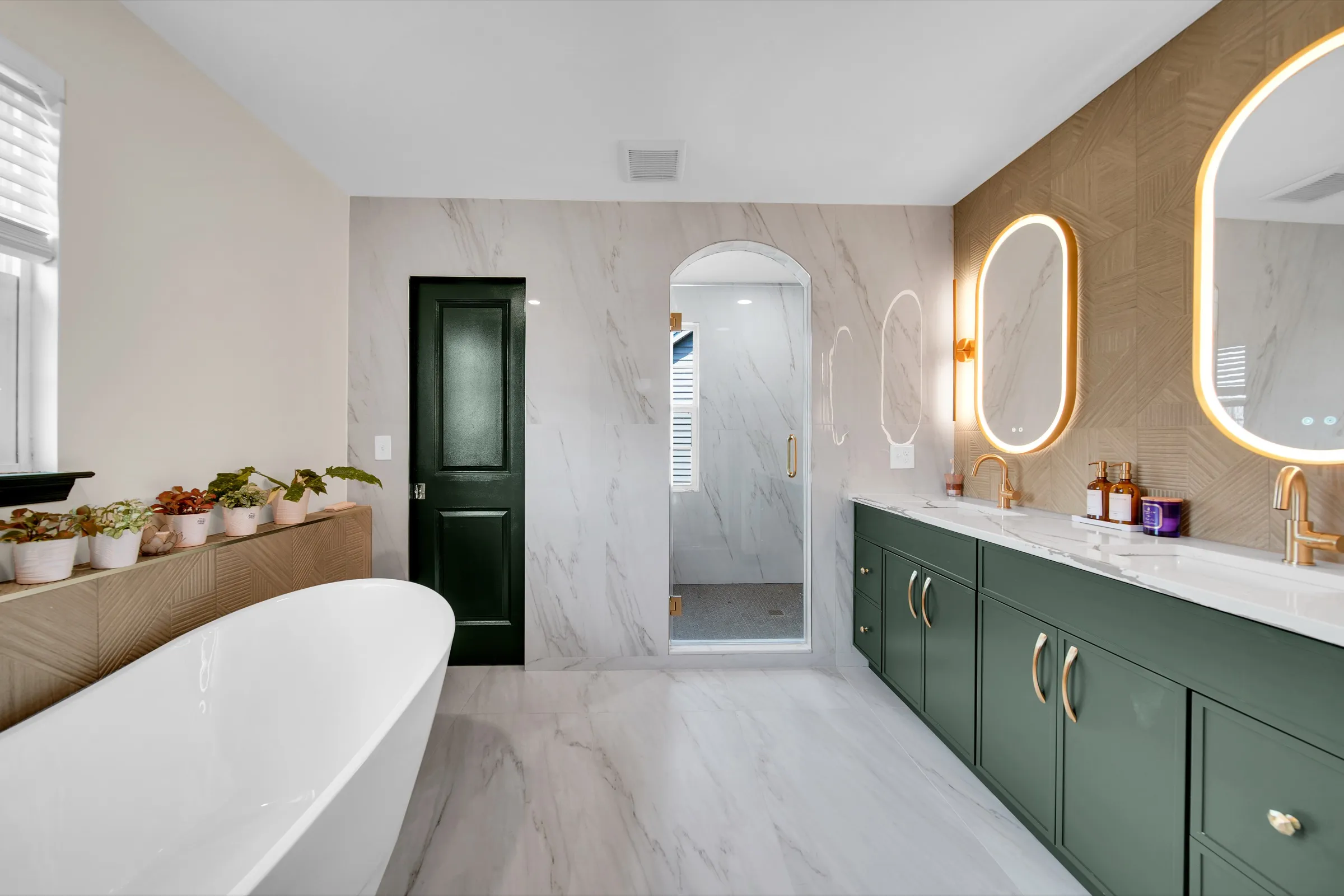 Remodeled primary bathroom with a freestanding tub, a hunter green double vanity, oval lit mirrors and gold fixtures