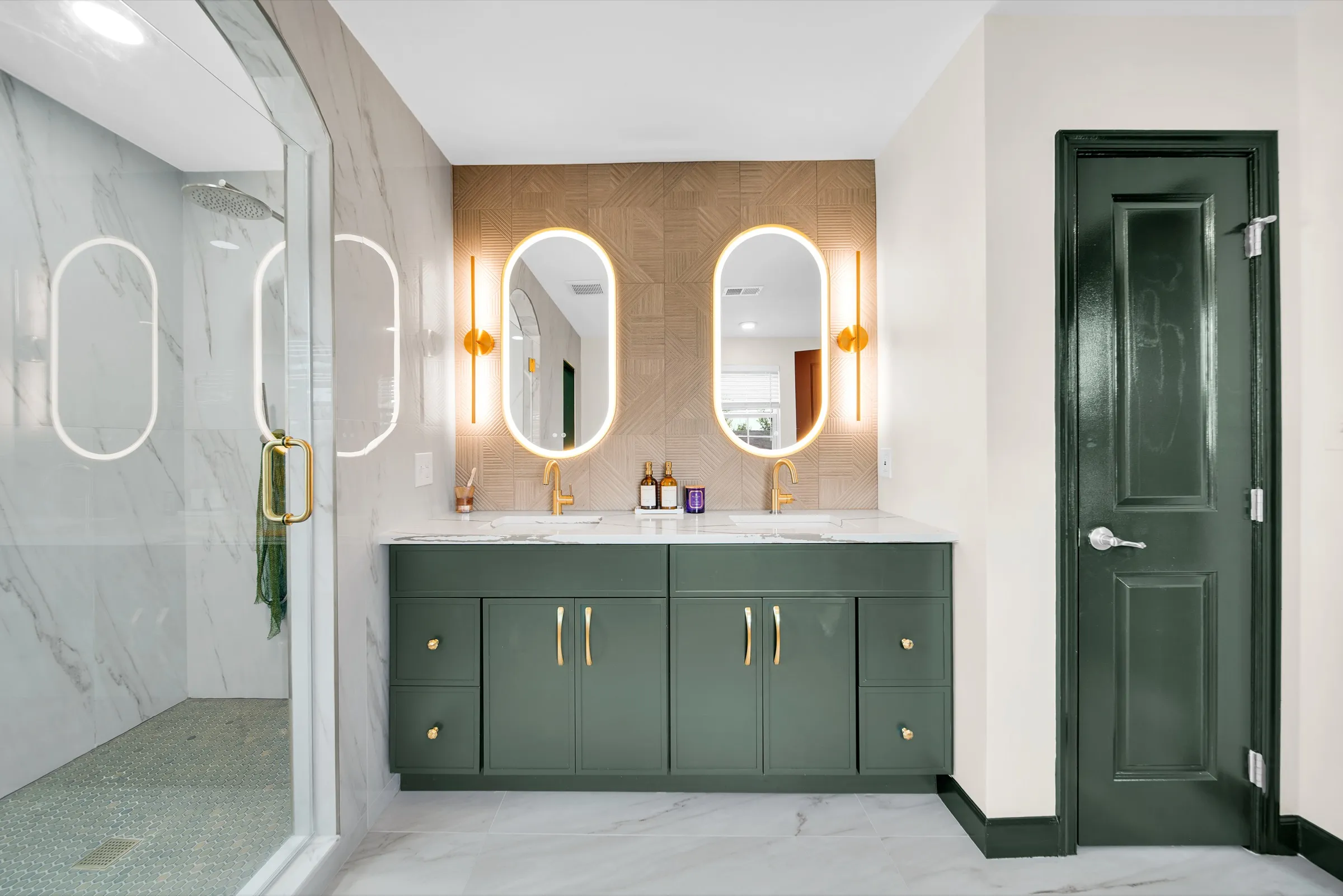 Hunter green double vanity with gold hardware, oval lit mirrors and a glass shower beside it